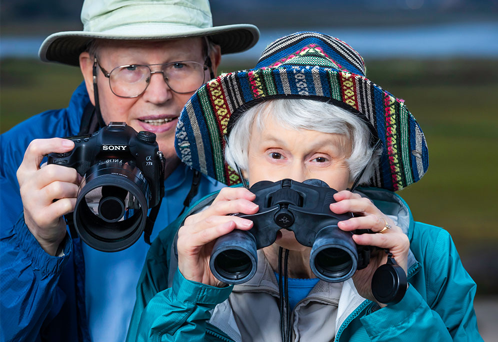 Bryce and Ann holding binoculars and digital camera outside in the wilderness